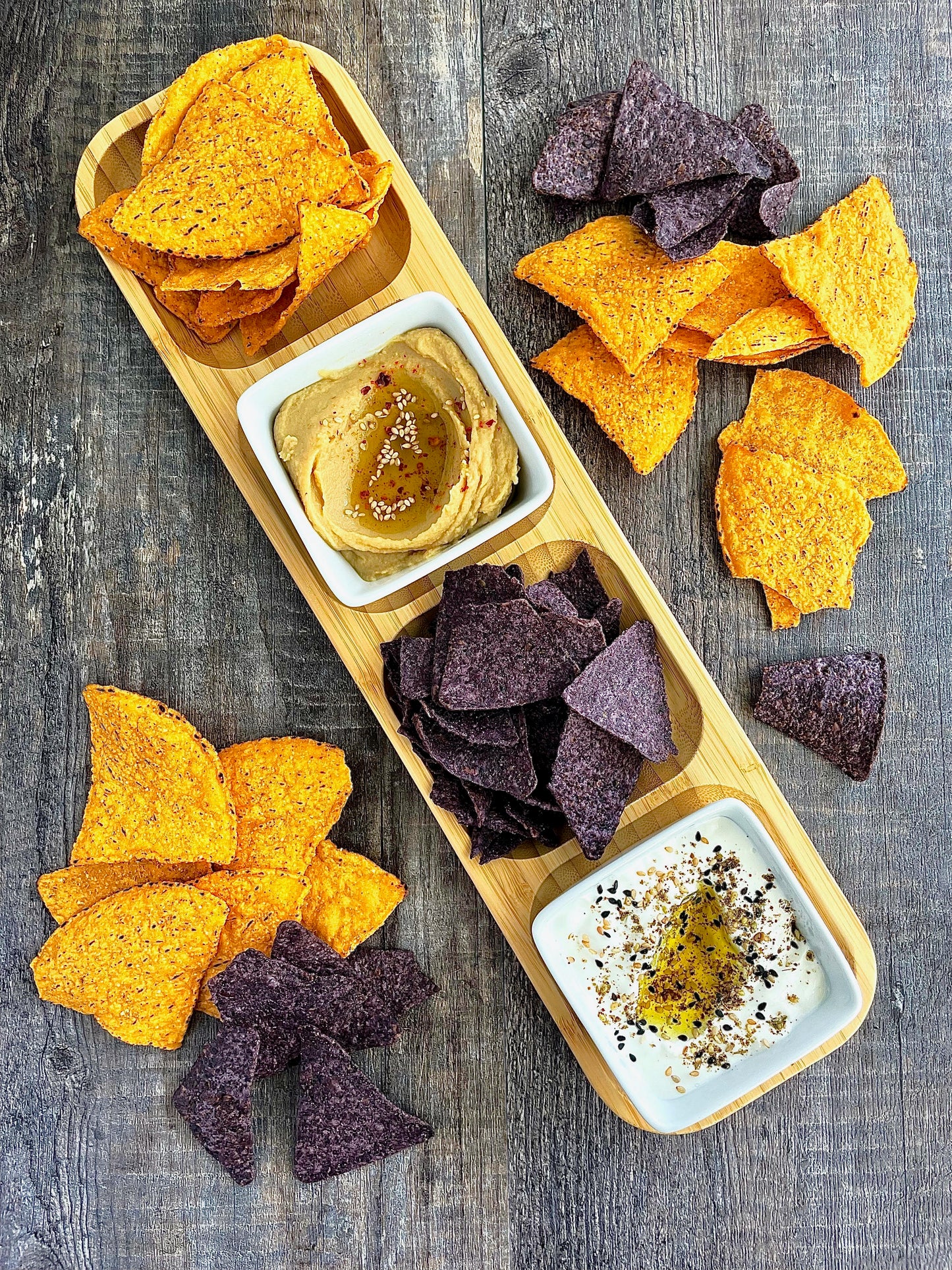 Bamboo serving board with yellow and purple tortilla chips, hummus, and olive oil dip in white bowls