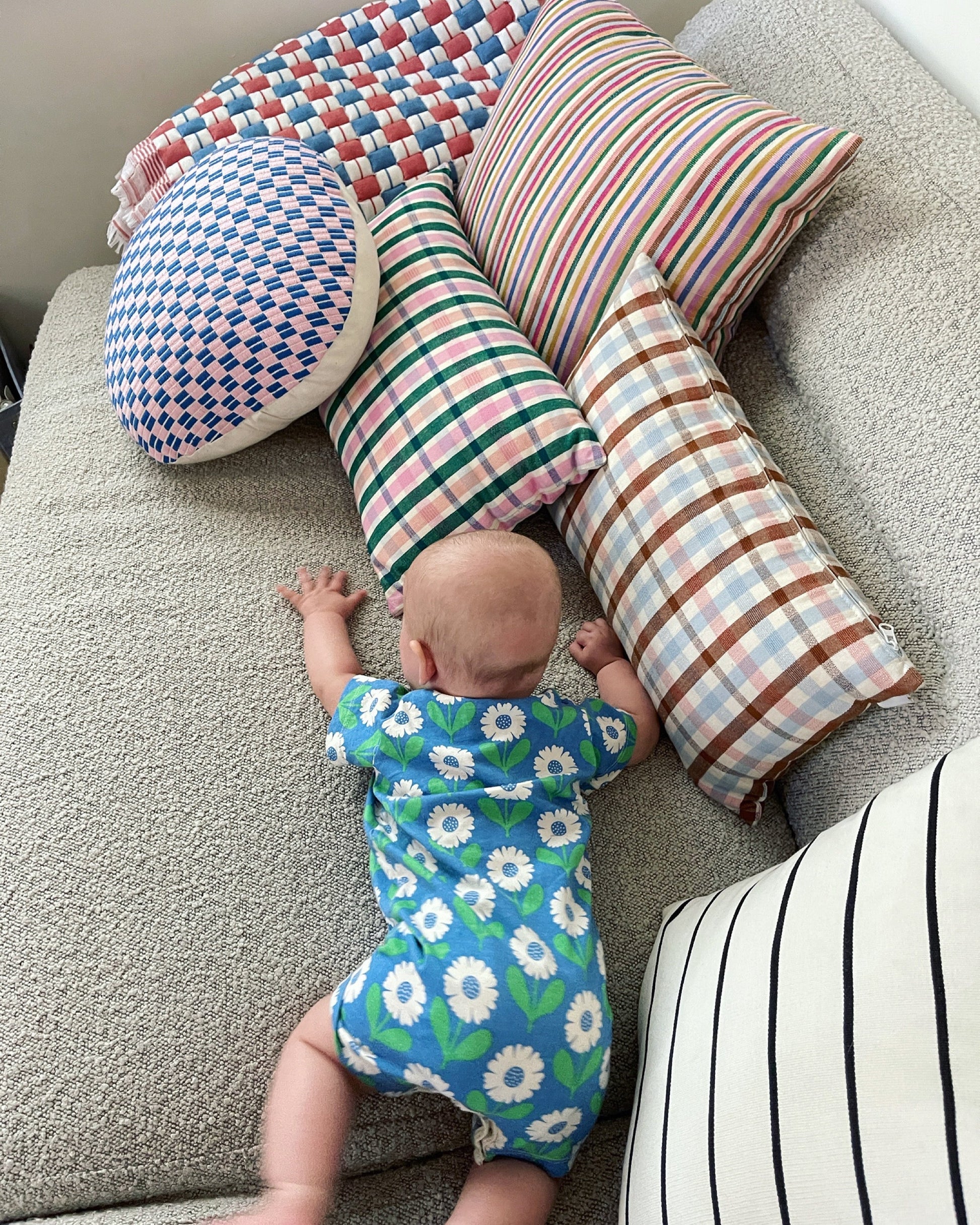 Colorful patterned pillows in striped, plaid, and geometric designs surrounding baby on gray carpet
