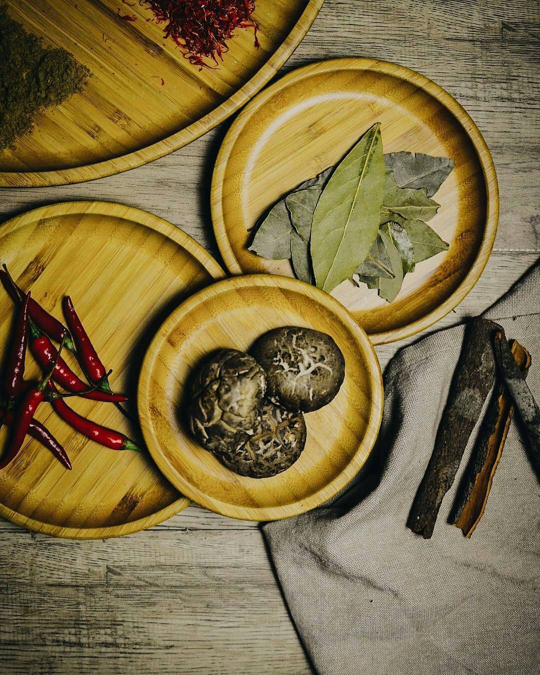 Bamboo round platter with natural grain texture, holding nuts and spices with red chilies and herbs nearby