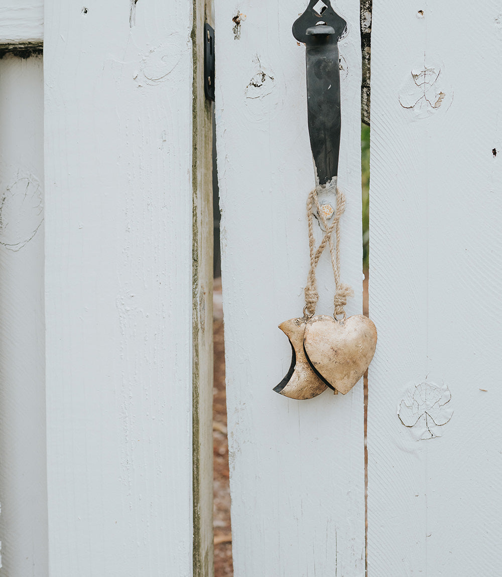 Rustic heart-shaped wind chime with brass bell and jute rope hanging against white wooden wall