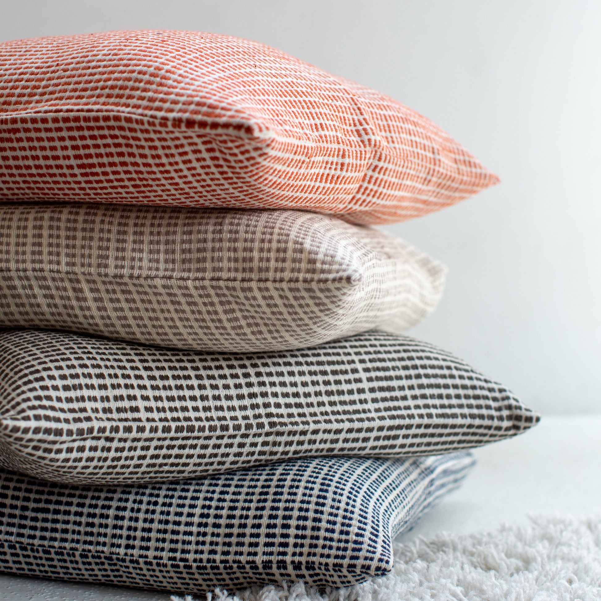 Stacked woven pillow covers in coral, taupe, charcoal, and blue with textured geometric patterns