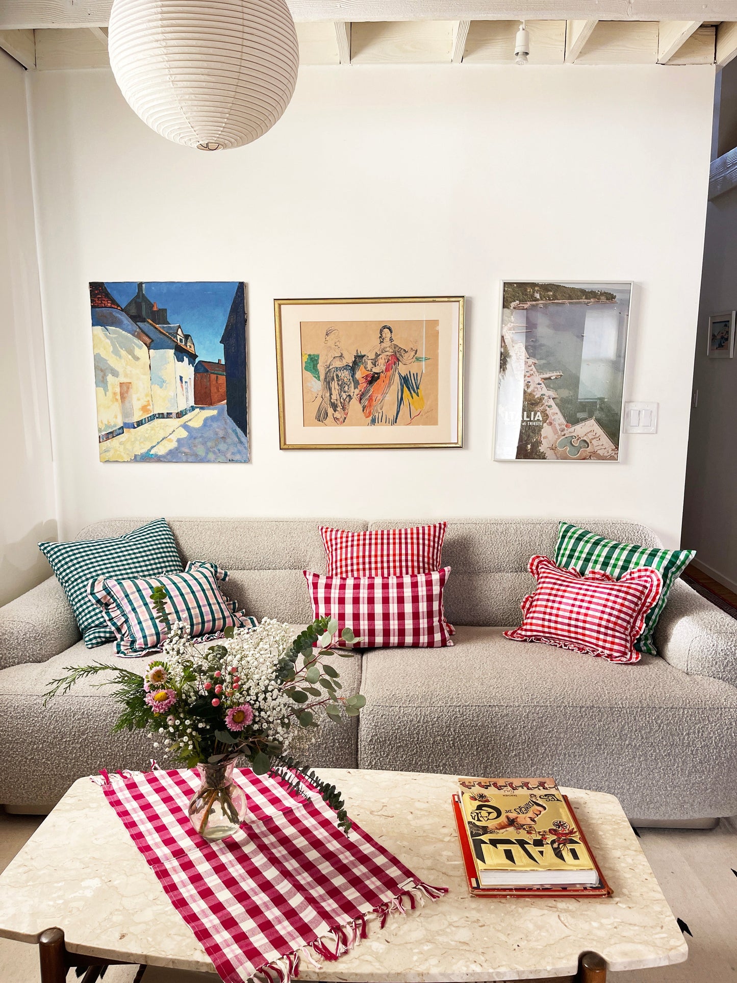 Red and white gingham plaid pillow with ruffle trim detailing, displayed on neutral linen sofa with coordinating striped pillows