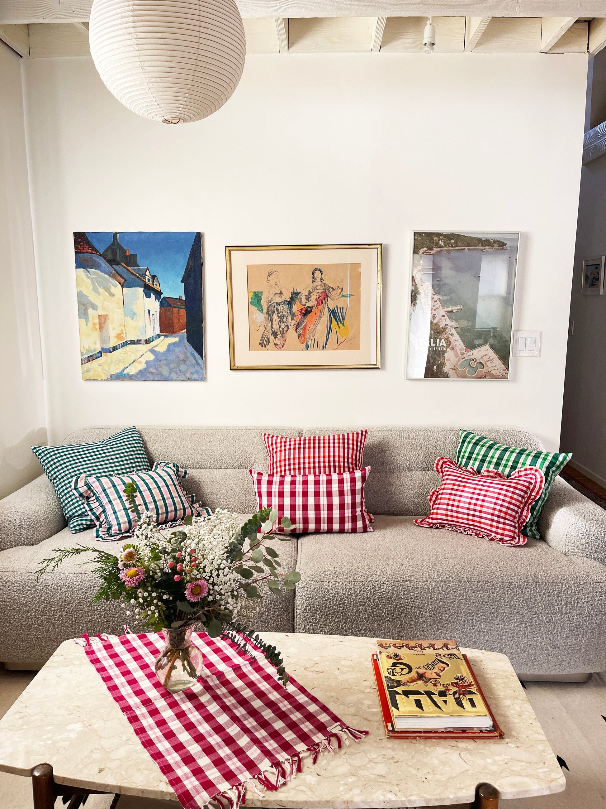 Red and white gingham plaid pillow with ruffle trim detailing, displayed on neutral linen sofa with coordinating striped pillows