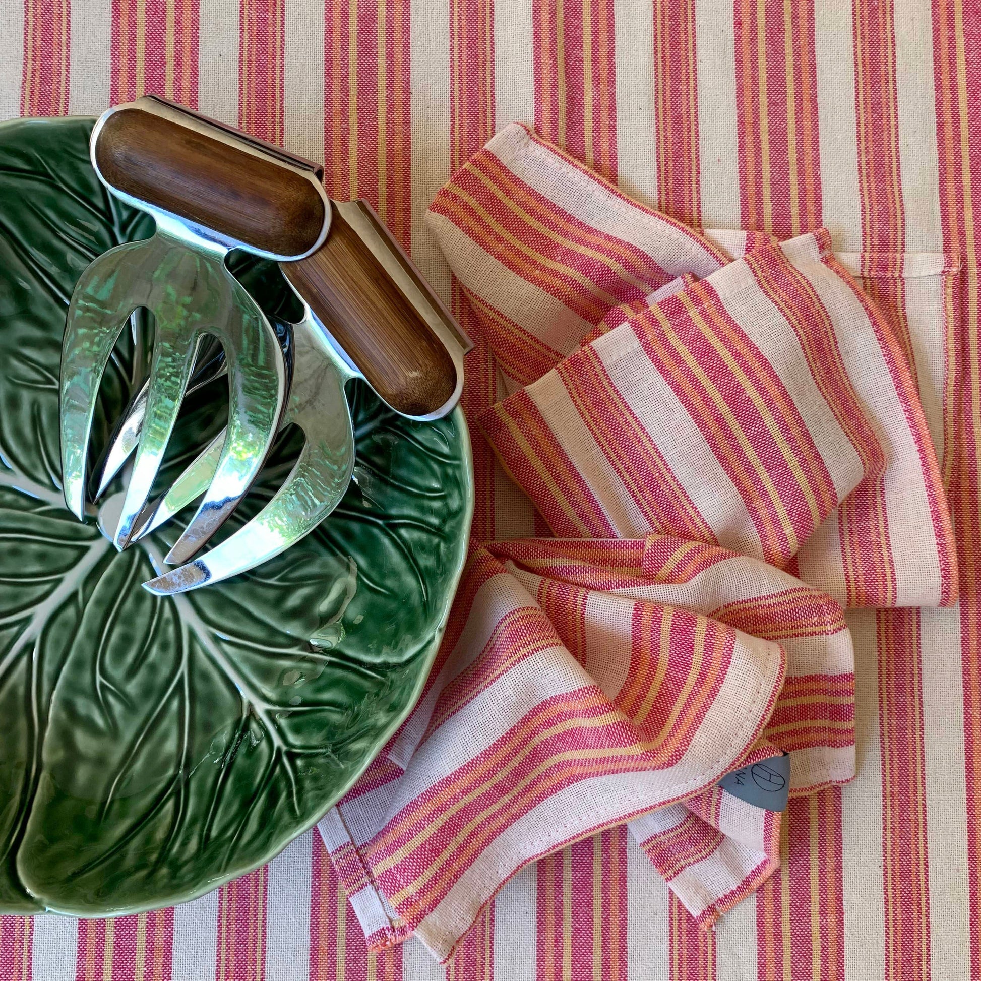 Magenta and cream striped linen tablecloth with matching napkins, styled with green ceramic leaf plate and wooden serving utensils