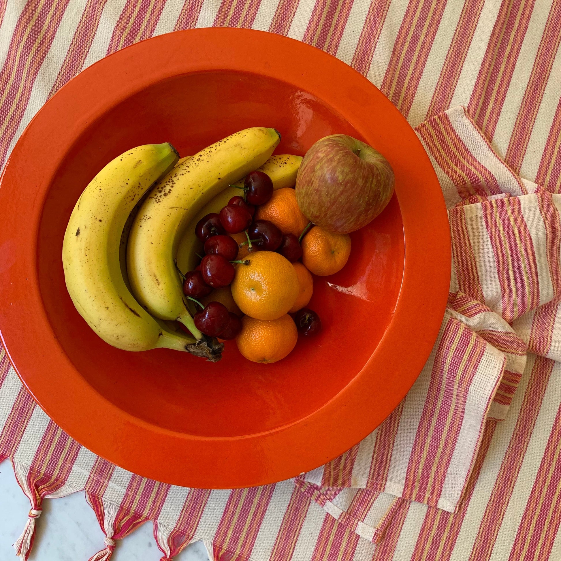 Bright red sustainable bowl filled with fresh bananas, cherries, oranges and apple on striped linen tablecloth
