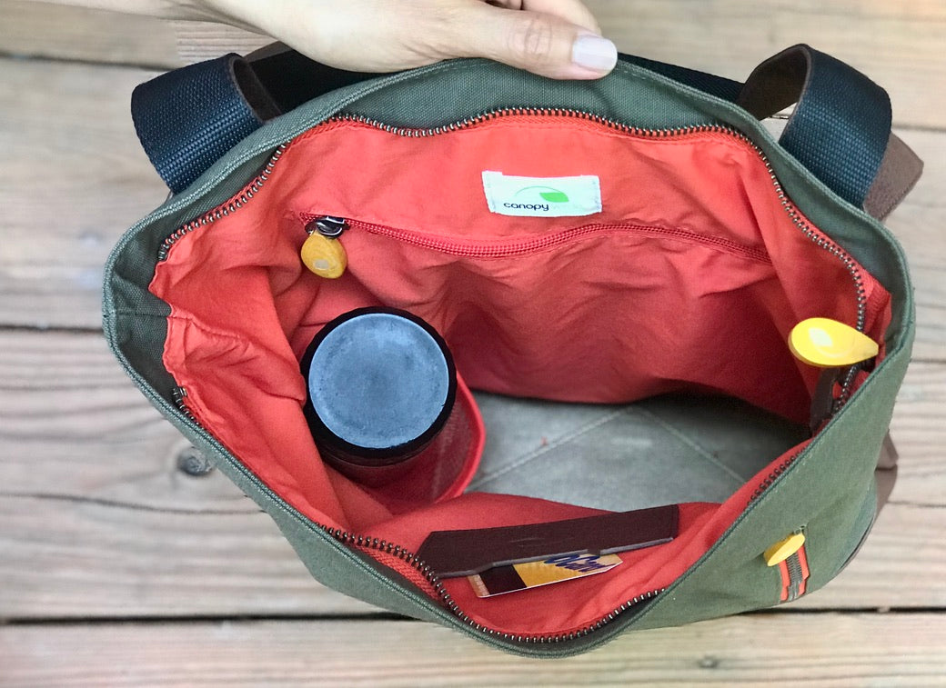 Sage green canvas tote backpack with red interior, navy straps, and multiple pockets, shown open with items inside