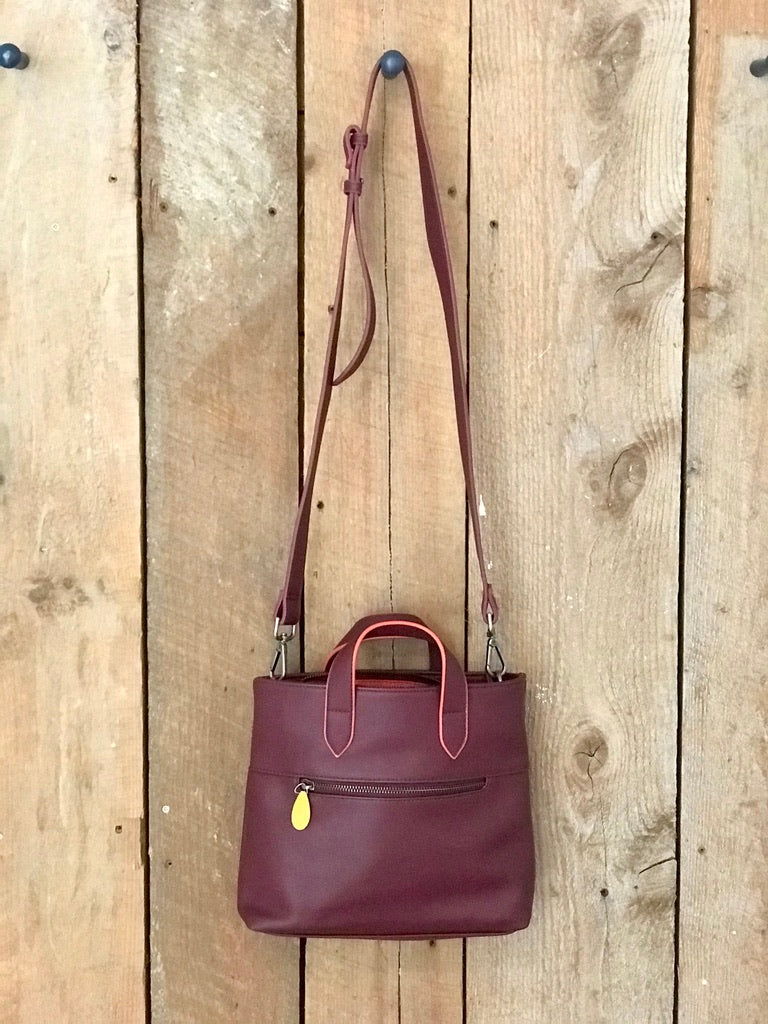 Burgundy leather crossbody satchel with dual handles, adjustable strap, and gold accent hardware on wooden background