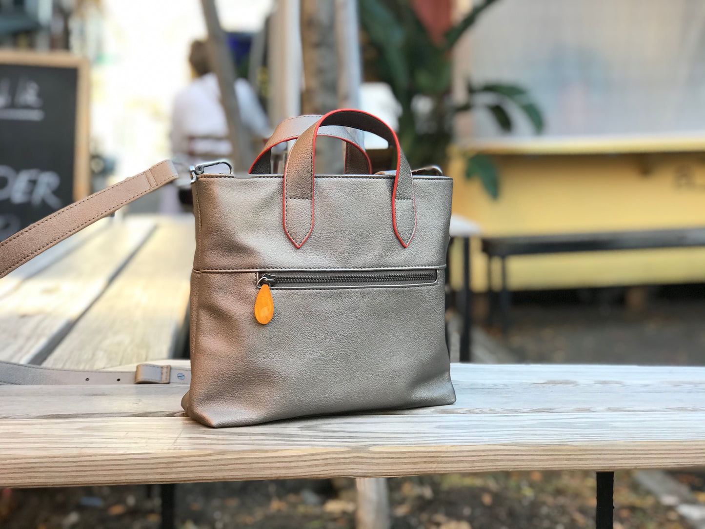 Beige leather crossbody satchel with red handles, zip pocket, and orange accent detail on wooden bench