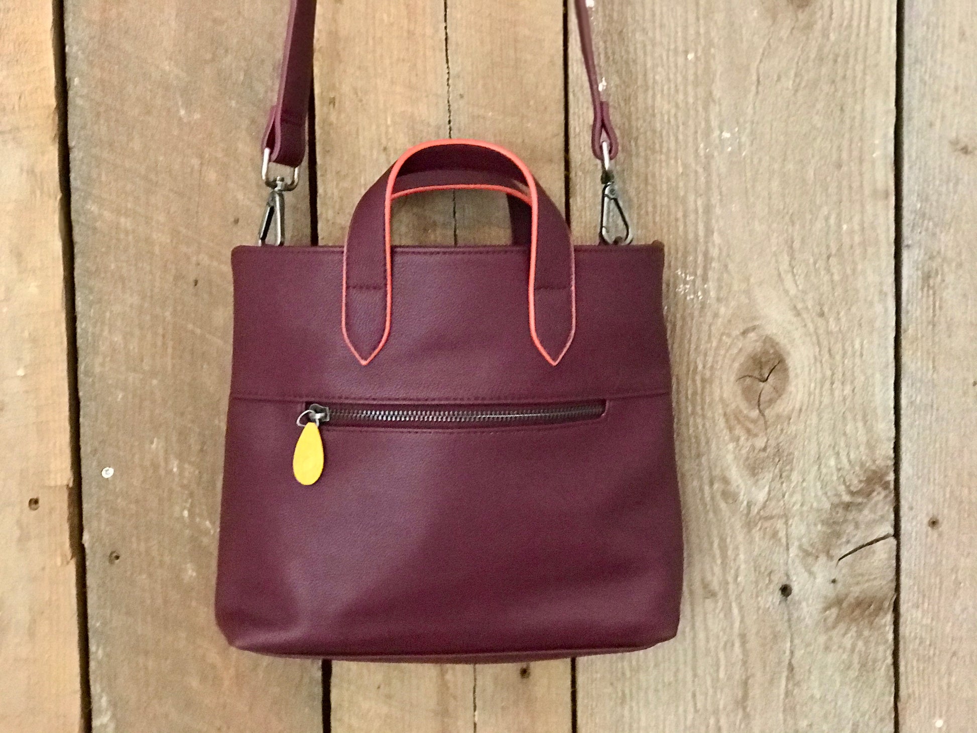 Burgundy leather crossbody satchel with coral-trimmed handles and yellow zipper accent, sustainable bag