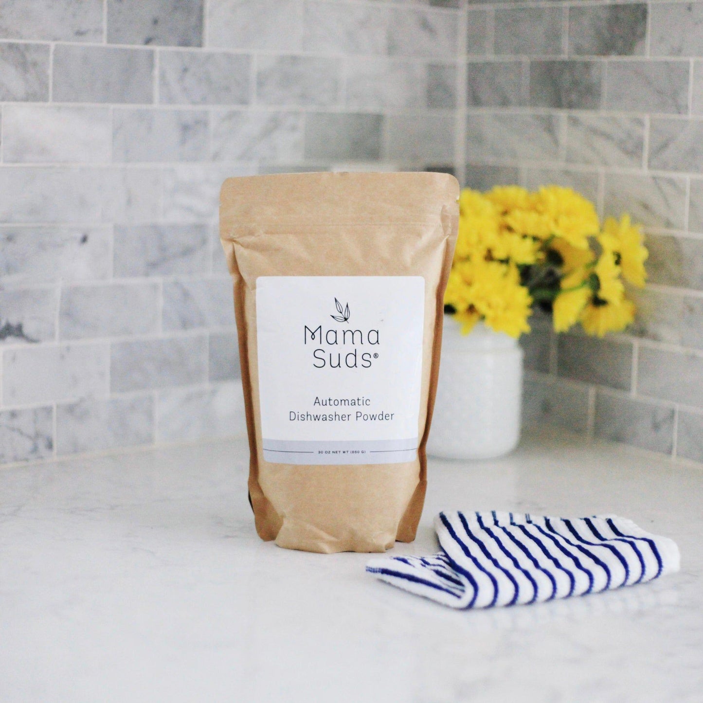 Kraft paper pouch of automatic dishwasher powder with white label, displayed with striped cloth and yellow flowers against marble tile