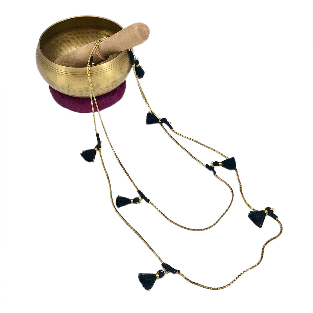 Brass singing bowl with wooden mallet, purple cushion, and gold chain necklace featuring black tassel details